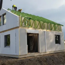 Wznoszenie dwukondygnacyjnego domu z prefabrykatów: montaż elementów konstrukcji dachu przez dwóch pracowników w żółtych kaskach, widoczny dźwig i krajobraz polny w tle.