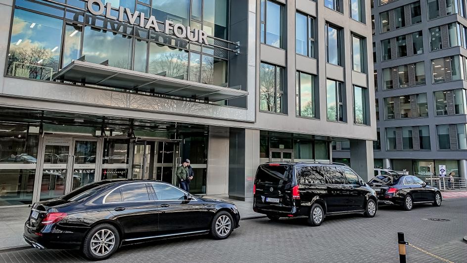 Czarne limuzyny i van Mercedes zaparkowane przed nowoczesnym budynkiem biurowym Olivia Four, sugerujące usługi transportu VIP.