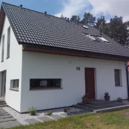 Nowoczesny, biały dom z ciemnoszarą dachówką, numerem 19 nad drzwiami i ścieżką z szarych płyt na białym żwirze.