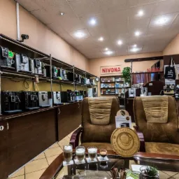 Salon z ekspresami do kawy: rzędy ekspresów na półkach, dwa brązowe fotele i szklany stolik z trzema kawami latte i jednym espresso.