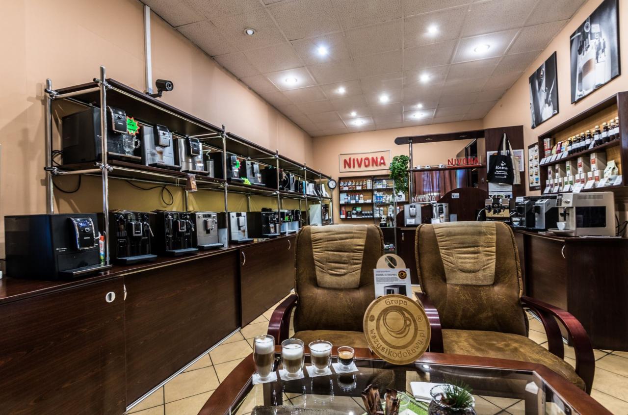 Salon z ekspresami do kawy: rzędy ekspresów na półkach, dwa brązowe fotele i szklany stolik z trzema kawami latte i jednym espresso.