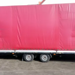 Czerwona plandeka naczepy ciężarowej z widocznymi mocowaniami i dwoma osiami kół, stojąca na ziemi