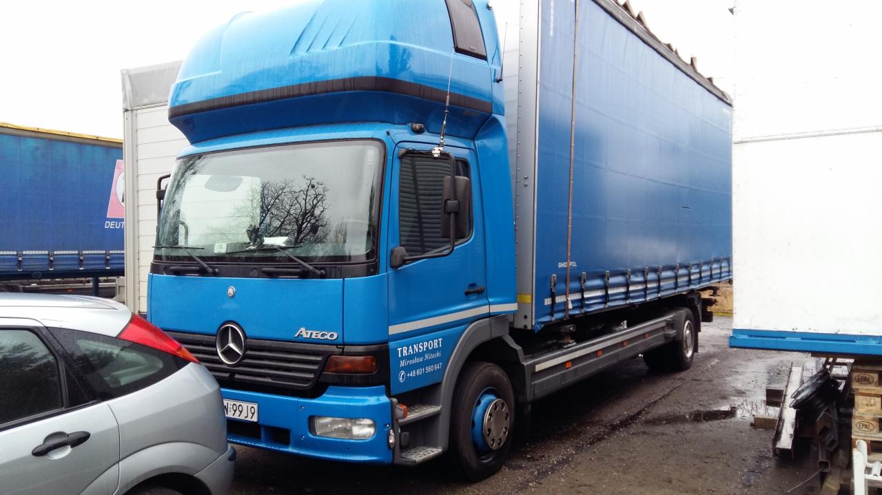 Niebieski samochód ciężarowy Mercedes Atego z plandeką, zaparkowany na placu, z widocznymi detalami konstrukcyjnymi i oznaczeniami firmy transportowej.