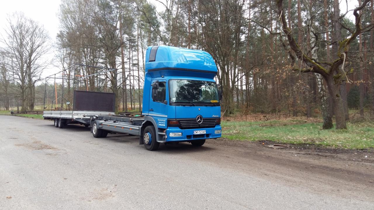 Niebieska ciężarówka Mercedes-Benz Actros z przyczepą platformową, częściowo załadowaną czarnym panelem, zaparkowana na asfaltowej drodze w pobliżu lasu w pochmurny dzień.