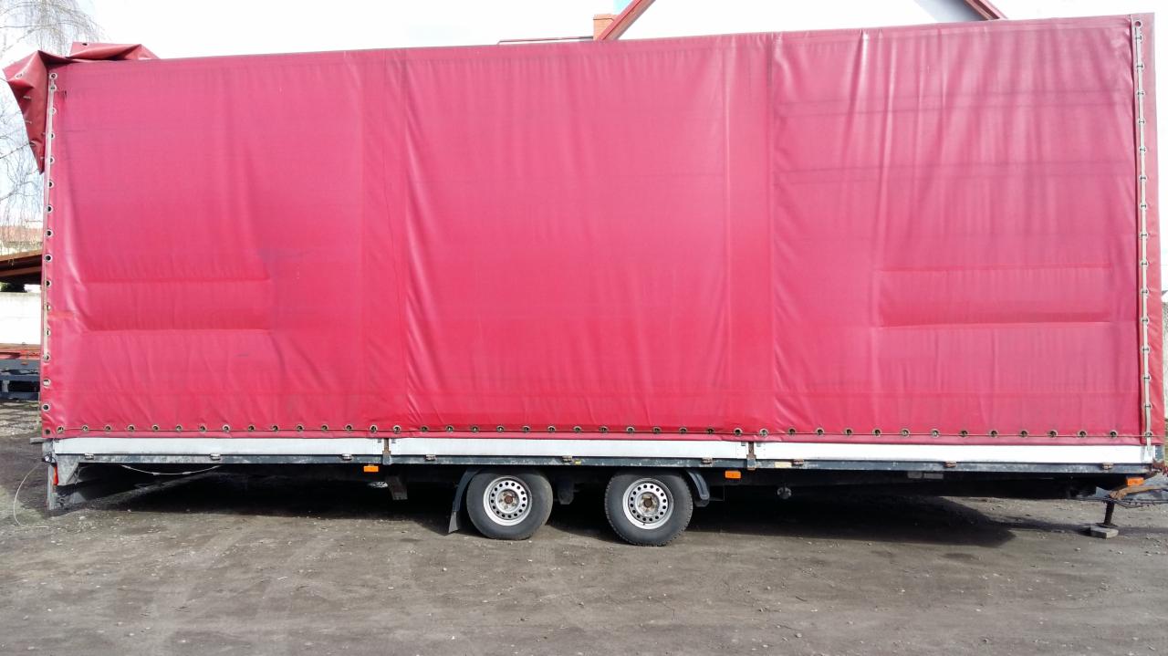 Czerwona plandeka naczepy ciężarowej z widocznymi mocowaniami i dwoma osiami kół, stojąca na ziemi