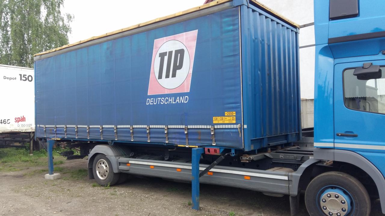 Niebieska naczepa ciężarowa z plandeką i logo TIP Deutschland, zaparkowana na żwirowym terenie, widoczny fragment kabiny ciągnika w kolorze błękitnym.