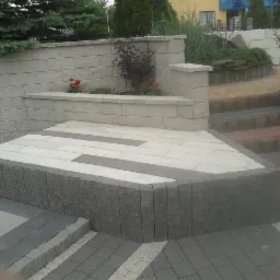 Kaskadowy taras z kostki brukowej w odcieniach szarości i bieli, z elementami zieleni w tle, prowadzący schodami do ogrodu.