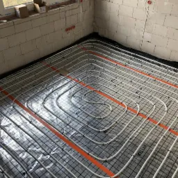 Białe rury ogrzewania podłogowego ułożone na czarnej macie w surowym wnętrzu z oknem i niezabudowanymi gniazdkami elektrycznymi.