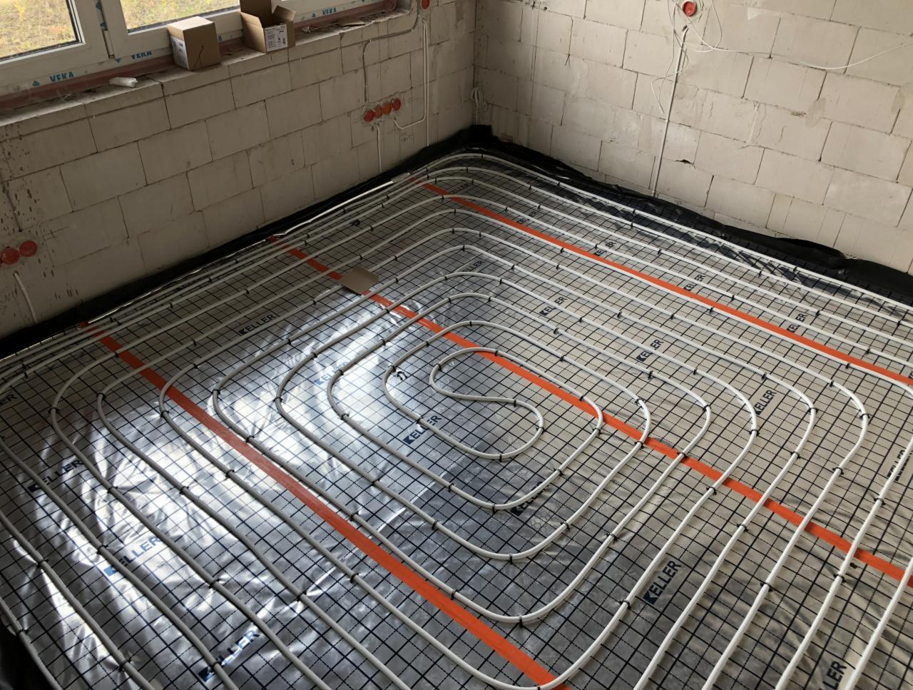 Białe rury ogrzewania podłogowego ułożone na czarnej macie w surowym wnętrzu z oknem i niezabudowanymi gniazdkami elektrycznymi.