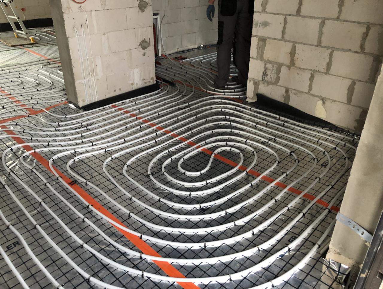 Białe rury systemu ogrzewania podłogowego ułożone w pętle na czarnej siatce z pomarańczowymi pasami, widoczne surowe ściany z betonu komórkowego i fragment sylwetki instalatora w rękawicach.