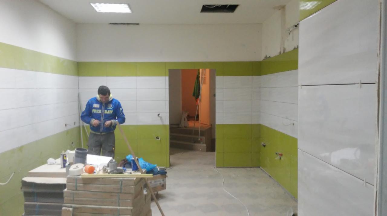 Pracownik w niebieskiej bluzie podczas układania białych płytek na ścianie łazienki, z widocznymi zielonymi pasami na dole i górze ściany, oraz narzędziami na prowizorycznym stole.