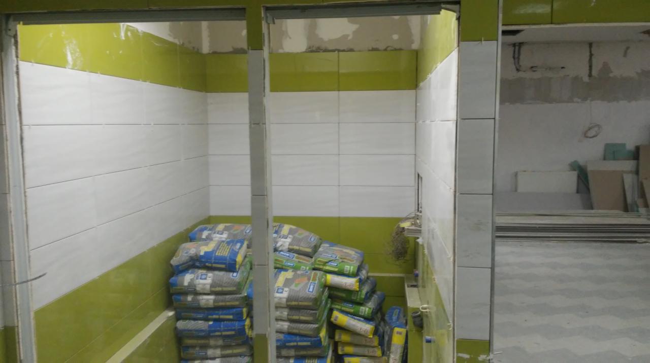 Wnętrze łazienki w trakcie remontu, z ułożonymi białymi płytkami i zielonym pasem na ścianach, worki z zaprawą ułożone na podłodze w brodziku, widoczne niedokończone prace w sąsiednim pomieszczeniu.