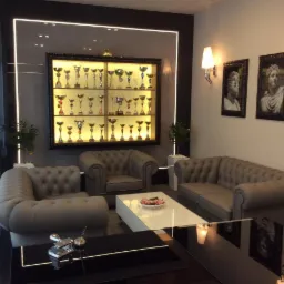 Eleganckie wnętrze biura z szarymi sofami typu chesterfield, białym stolikiem kawowym, ścianą z podświetlaną gablotą wypełnioną trofeami i portretami w ramach na ścianie.