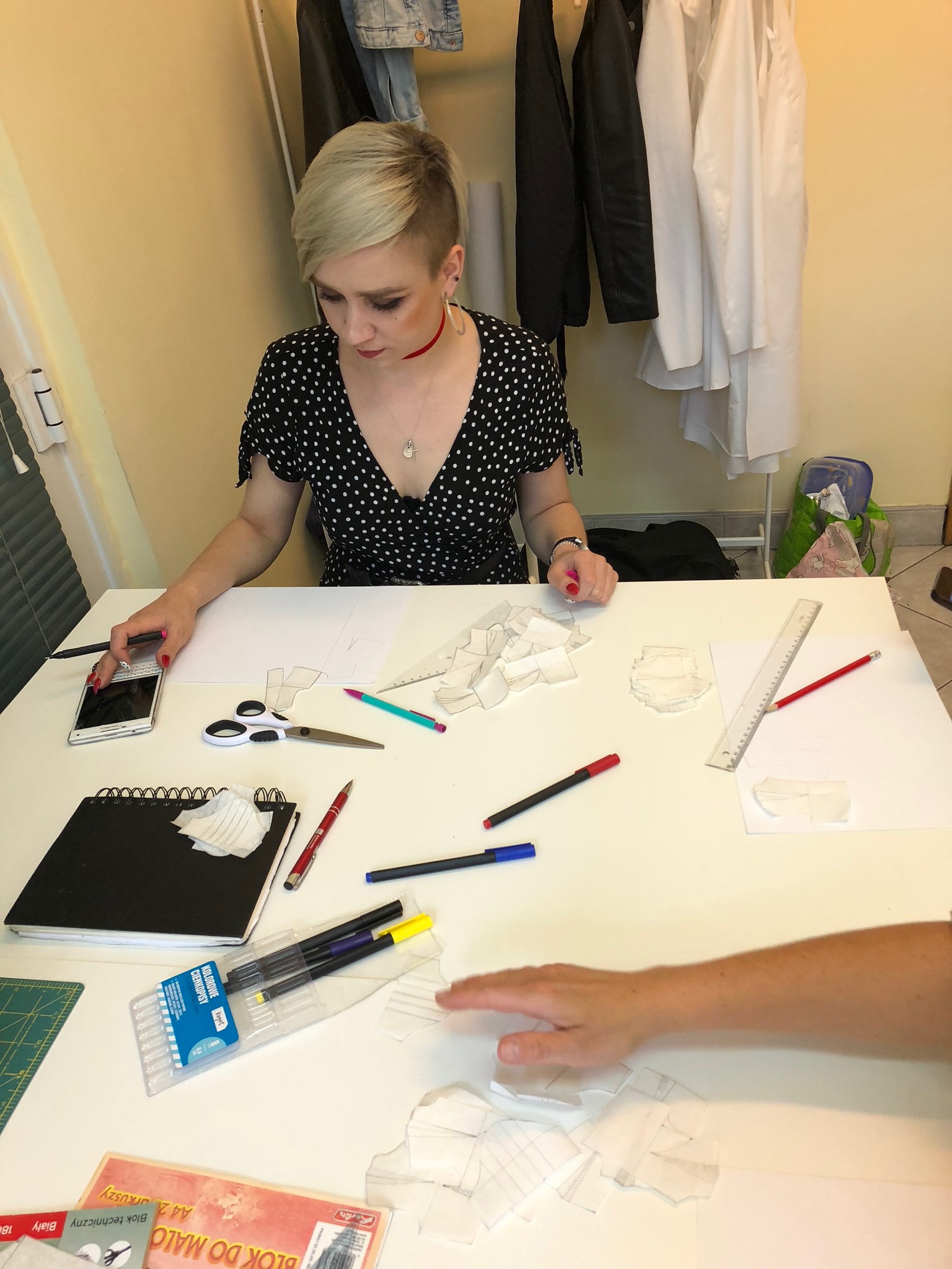 Kobieta z krótkimi blond włosami w czarnej sukience w białe kropki, pochylona nad białym stołem z rozłożonymi szablonami, notatnikiem, nożyczkami, długopisami i telefonem komórkowym, prawdopodobnie...