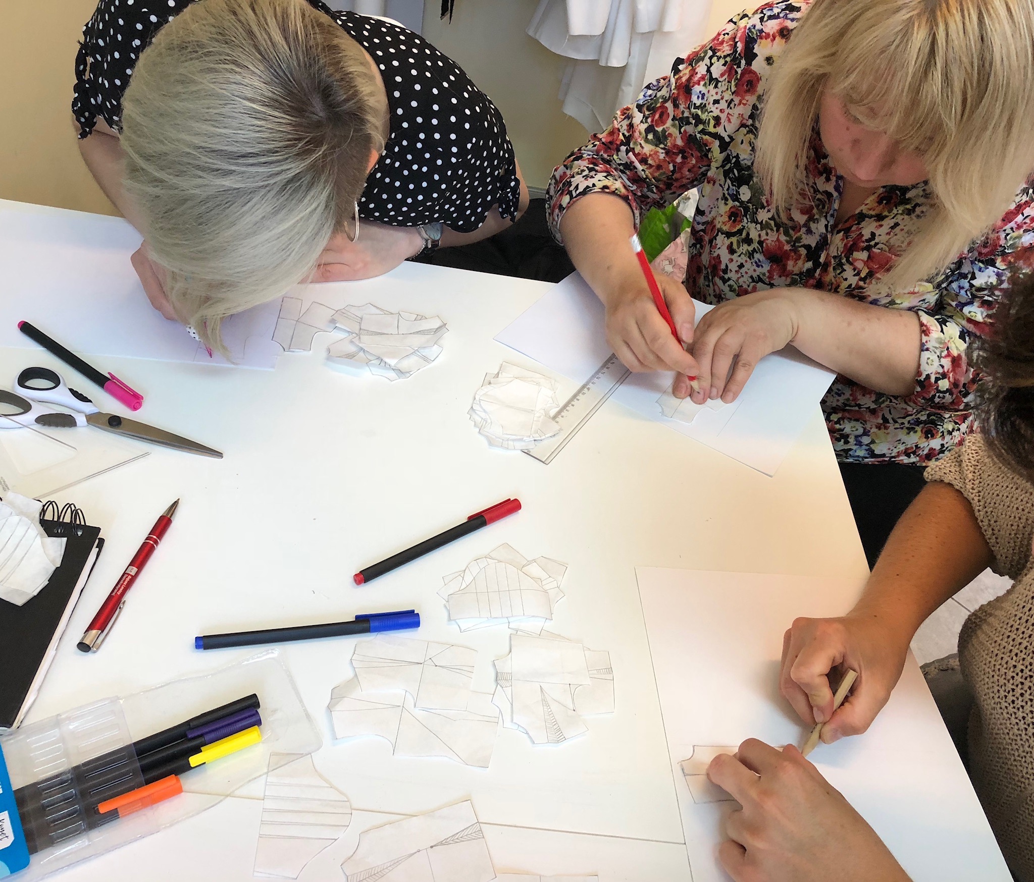 Uczestnicy warsztatów kreślą szablony odzieży na białym stole, używając ołówków, linijek i nożyczek; widoczne pocięte fragmenty szablonów, mazaki i notatnik.