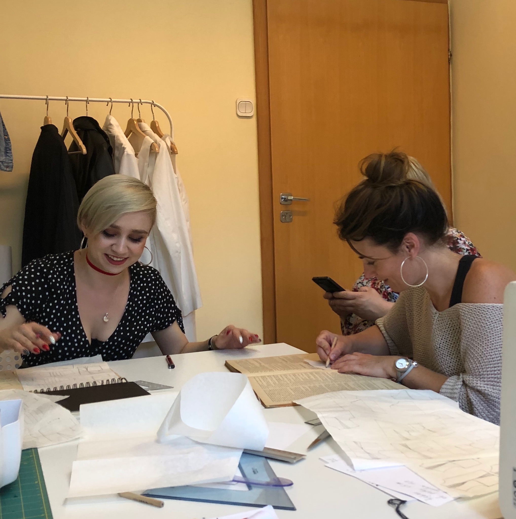Dwie kobiety pracują nad projektami odzieży przy stole: blondynka w czarnej sukience w białe kropki z czerwonym chokerem i brunetka w swetrze, jedna z nich korzysta z telefonu komórkowego.