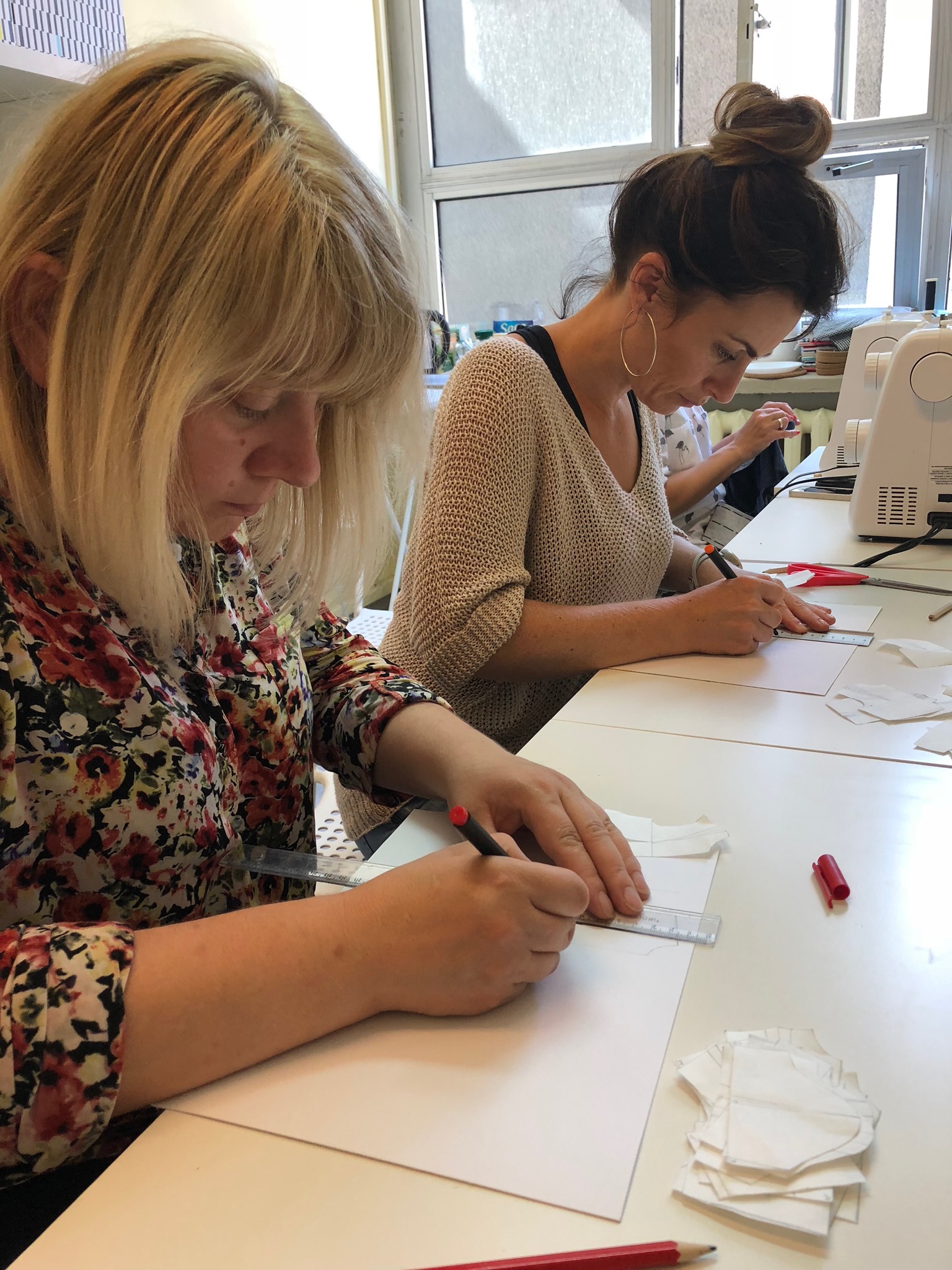 Dwie kobiety przy białych stołach rysują szablony na papierze, używając linijki i ołówka, w tle widoczne maszyny do szycia.