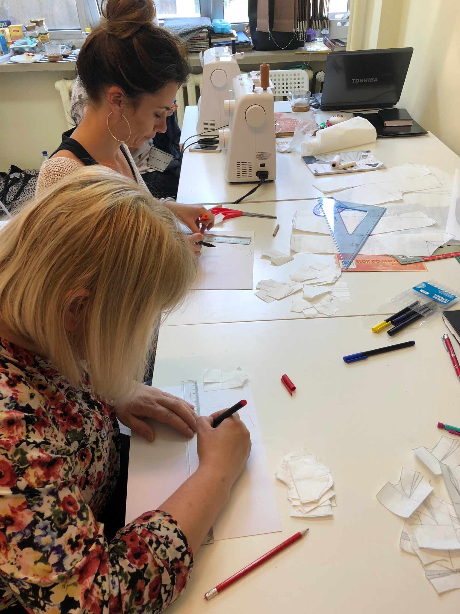 Dwie kobiety pracują nad projektami odzieży przy białym stole, używając linijek, ołówków i szablonów; w tle maszyna do szycia i laptop.