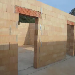 Wznoszenie ścian z bloczków ceramicznych z widocznymi otworami okiennymi i drzwiowymi, wzmocnionymi nadprożami z cegły kratówki, na surowym betonowym podłożu.