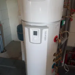 Biały, cylindryczny zasobnik ciepłej wody użytkowej marki Atlantic z panelem sterowania, widoczne przyłącza hydrauliczne i rury instalacji grzewczej w tle.