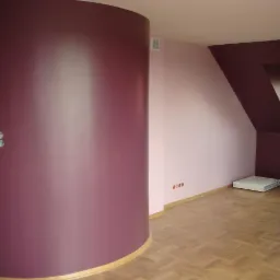 Pomalowane wnętrze z zaokrągloną ścianą w kolorze śliwki, jasnor&oacute;żową ścianą i ciemnofioletową ścianą ze skosem, parkiet na podłodze.