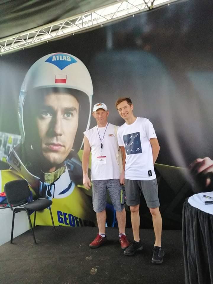Dwaj mężczyźni pozują na tle baneru z wizerunkiem sportowca w kasku Atlas, z polską flagą. Jeden z mężczyzn ma czapkę i identyfikator.