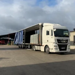 Ciężarówka MAN z otwartą naczepą, załadowana paletami z workami pelletu, na placu przed magazynem.