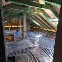 Instalacja ogrzewania podłogowego w surowym stanie poddasza, widoczne rozłożone rury na folii z siatką, drewniana konstrukcja dachu, narzędzia i wiadro w tle.