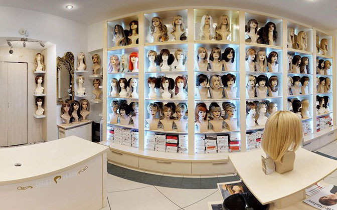 Wnętrze sklepu z perukami: ekspozycja na regałach z podświetleniem LED, manekiny z różnorodnymi fryzurami, blond peruka na pierwszym planie na blacie.