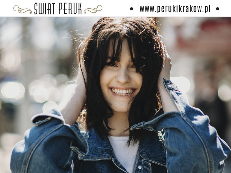 Uśmiechnięta młoda kobieta z ciemnymi włosami i grzywką, prezentująca perukę, w dżinsowej kurtce na tle rozmytego miejskiego otoczenia, logo 'Świat Peruk' i adres strony internetowej u góry.