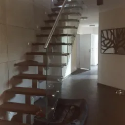 Nowoczesne schody z drewnianymi stopniami i szklaną balustradą w minimalistycznym wnętrzu, z betonową ścianą i legowiskiem dla psa u dołu.