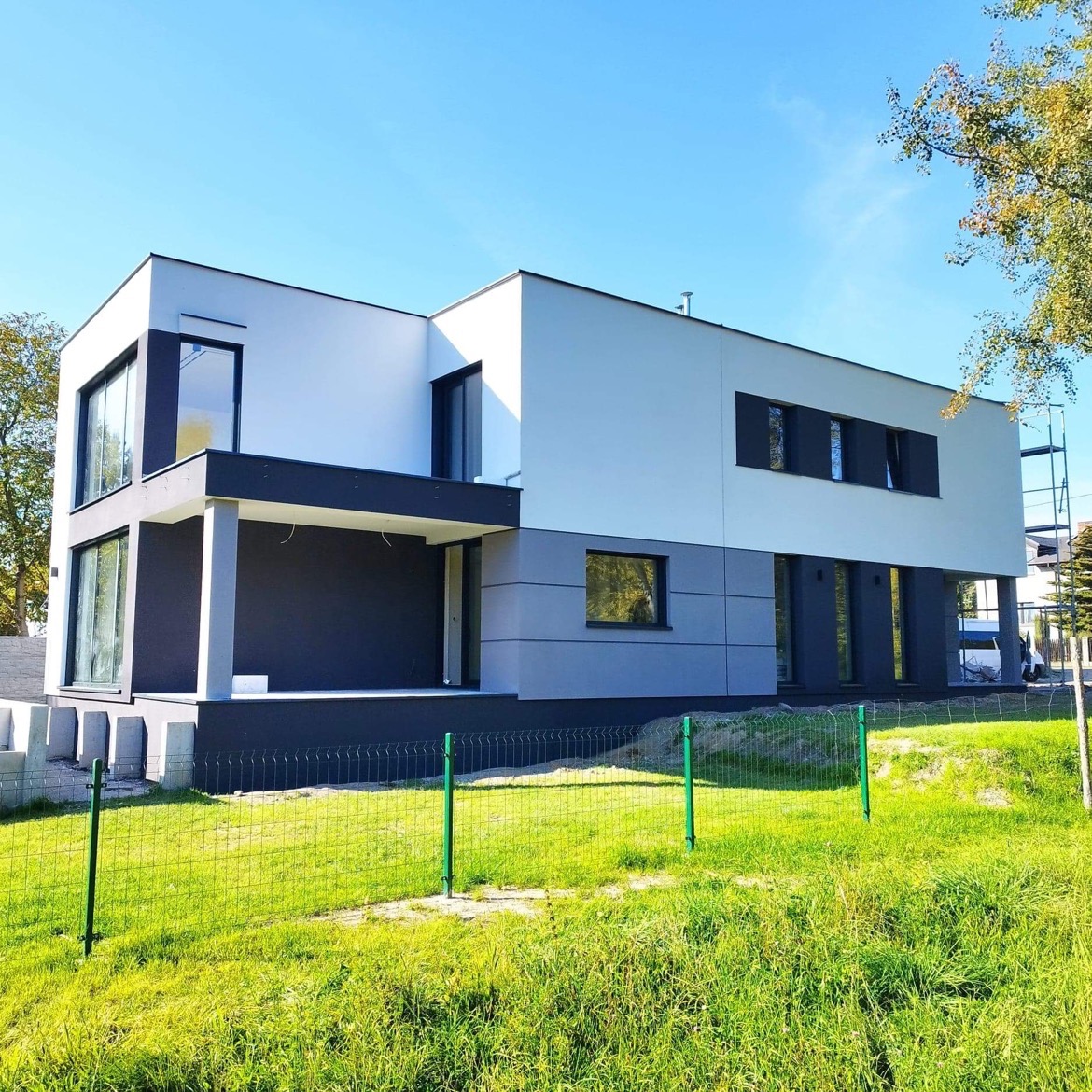 Nowoczesny, dwukondygnacyjny dom jednorodzinny w Katowicach z płaskim dachem, wykończony w bieli i szarości, z dużymi oknami i minimalistycznym balkonem, otoczony zielonym trawnikiem i ogrodzeniem...