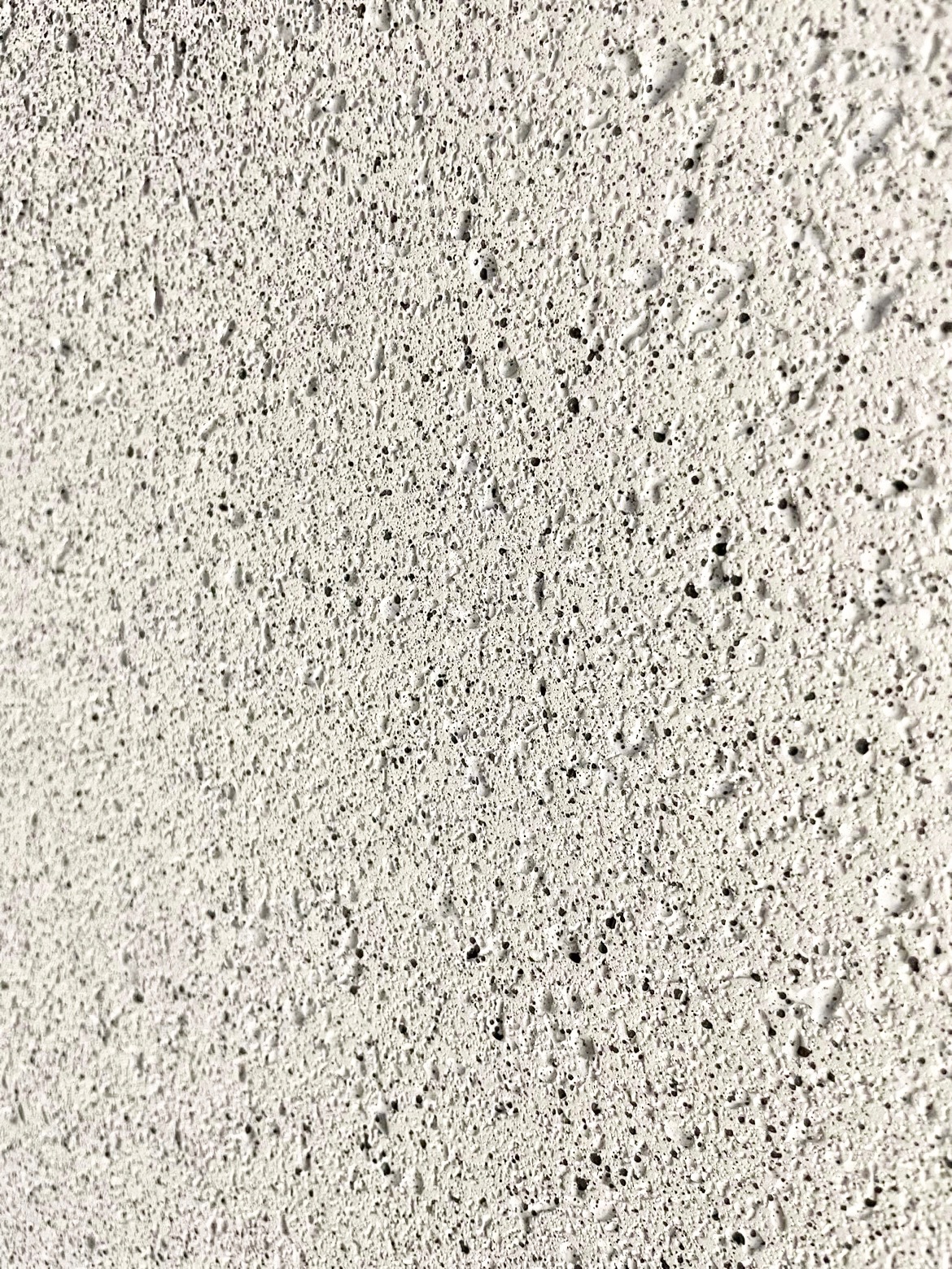 Zbliżenie na białą, porowatą ścianę z wyraźną fakturą i drobnymi otworami, oświetloną z boku, tworzącą delikatny cień.