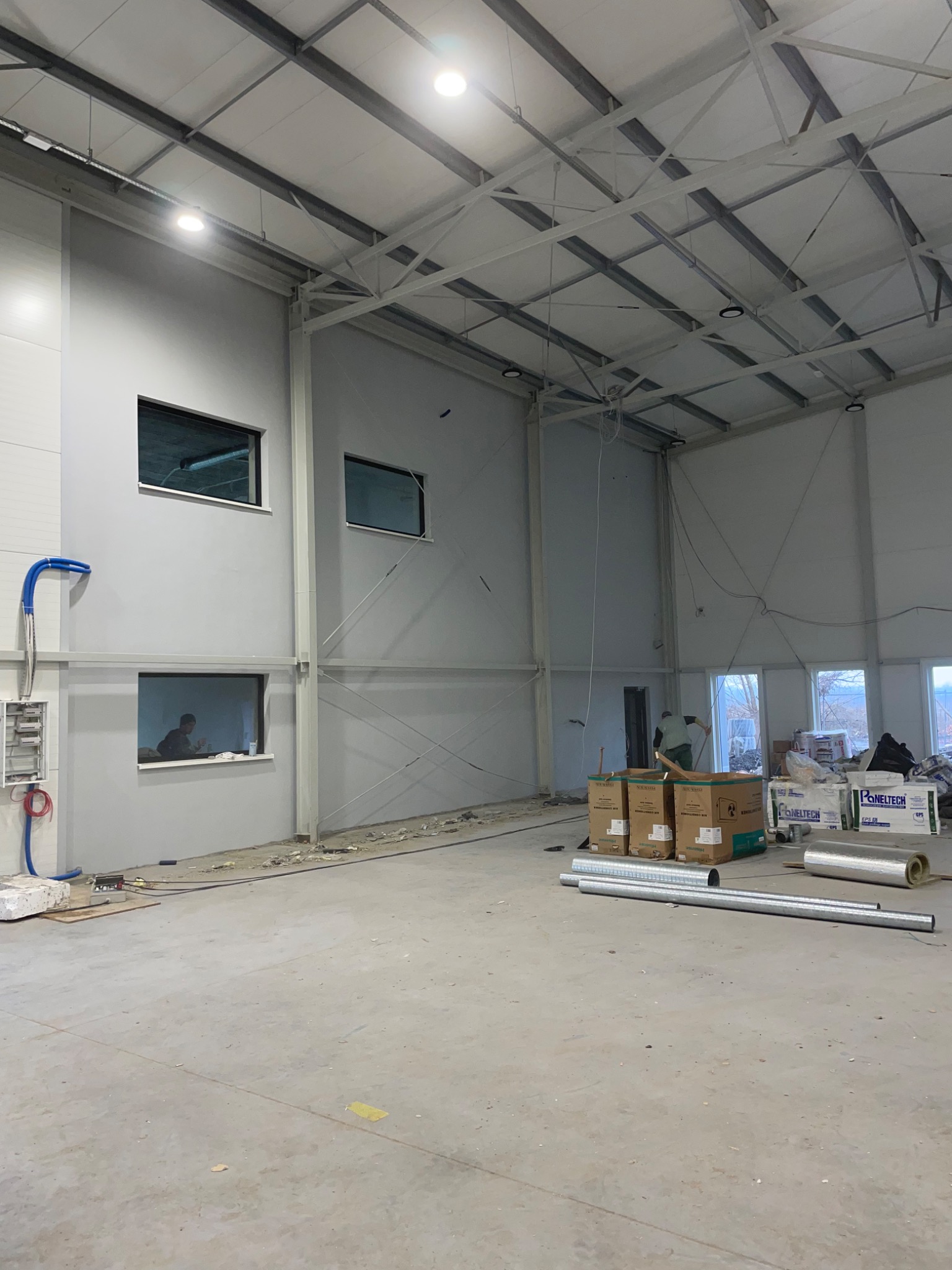 Wnętrze hali w trakcie wykończenia, widoczne materiały budowlane, rury wentylacyjne na podłodze, pracownik widoczny w oknie, sufit z widoczną konstrukcją stalową i oświetleniem.