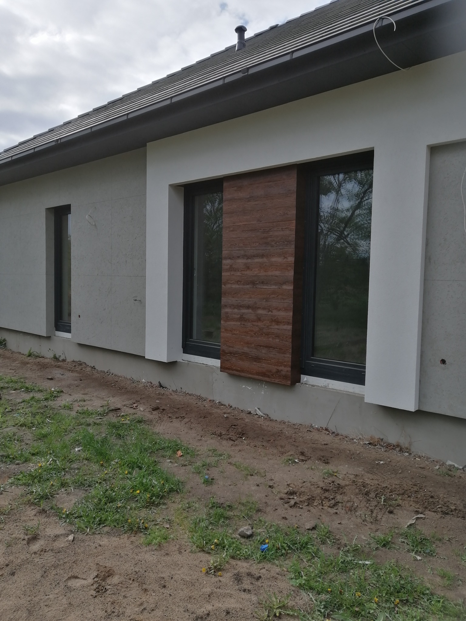 Elewacja budynku z oknami: szara płyta elewacyjna, białe obramowania okien, okno z brązową okładziną imitującą drewno, trawnik z ziemią w tle.
