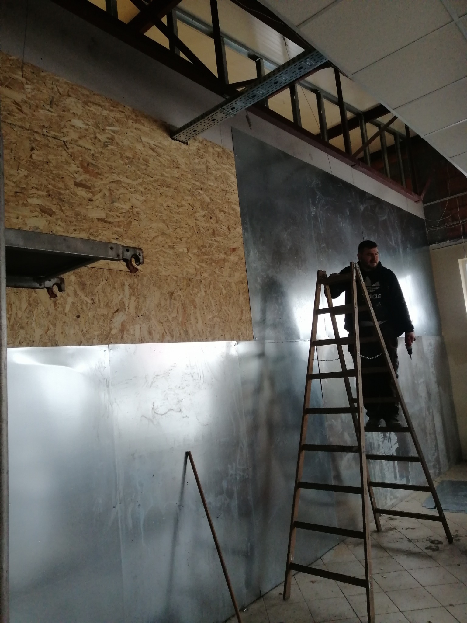 Pracownik na drabinie z wiertarką, montaż płyty OSB i blachy na ścianie w trakcie remontu wnętrza, widoczne elementy konstrukcyjne sufitu.