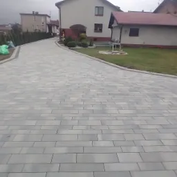 Ułożona szara kostka brukowa na podjeździe do domu jednorodzinnego, widoczna betonowa mieszarka i worki na trawniku po lewej stronie.