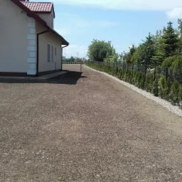 Przygotowany teren pod trawnik z obrzeżem z białego grysu i posadzonymi tujami wzdłuż ogrodzenia z siatki.