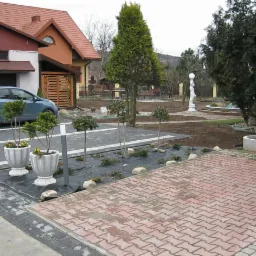 Nowo zaprojektowany ogród z kostką brukową, roślinami w donicach i małą architekturą, widoczny na tle domu i zaparkowanego samochodu.