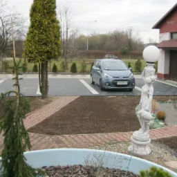 Nowo ułożona, grafitowa kostka brukowa na podjeździe przed domem z zaparkowanym, błękitnym samochodem, widoczna ozdobna lampa ogrodowa w kształcie rzeźby kobiety, trawnik w trakcie zakładania.