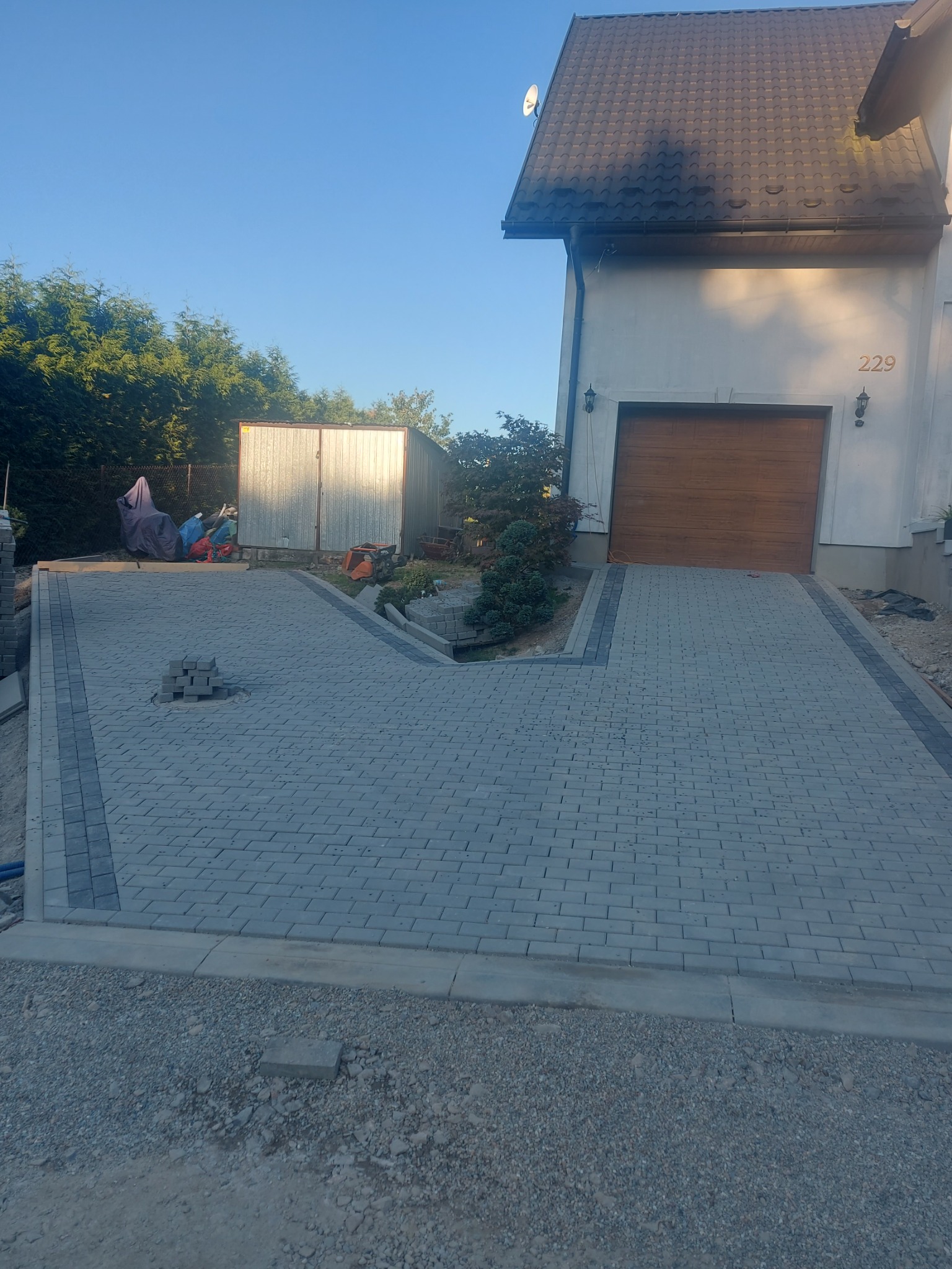 Nowa, szara kostka brukowa ułożona na podjeździe przed domem z brązową bramą garażową; widoczne obrzeża z ciemniejszej kostki, stos kostki na środku podjazdu i przygotowane podłoże.