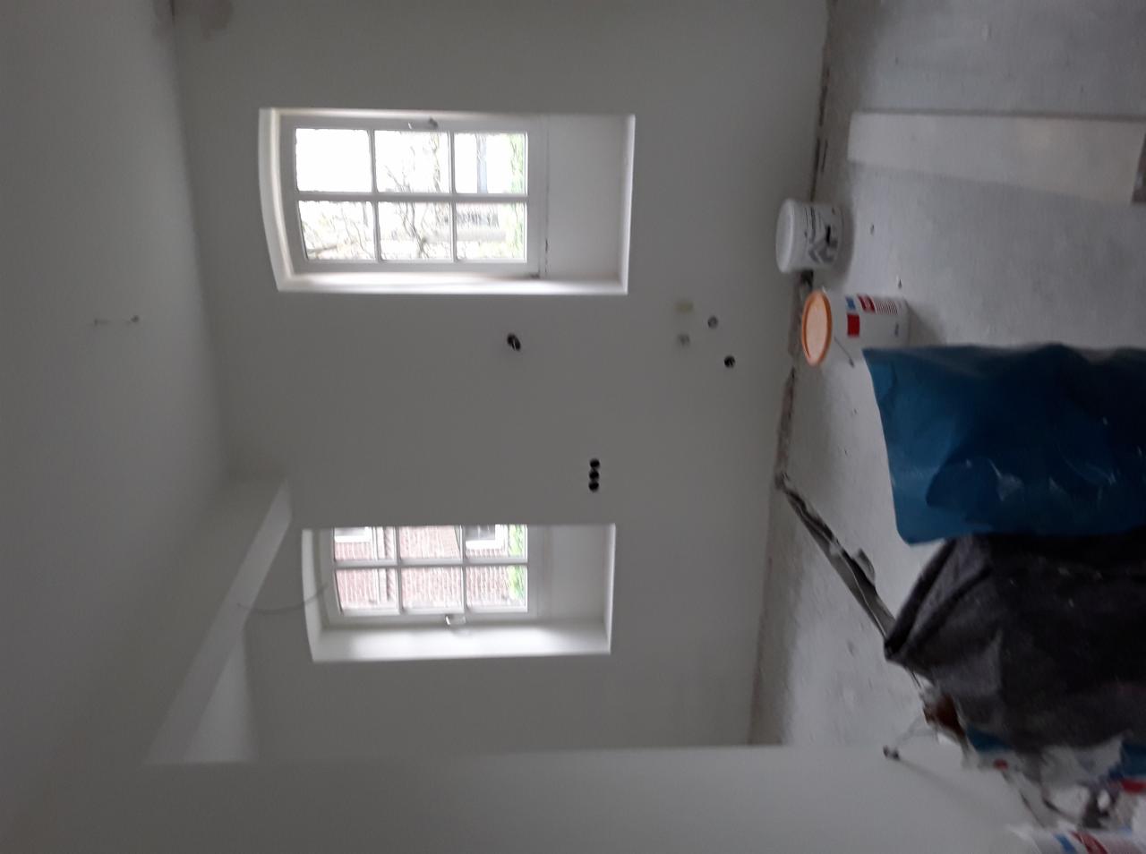 Wnętrze w trakcie malowania ścian na biało, widoczne dwa okna, puszki z farbą i zabezpieczona podłoga.