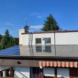 Panele słoneczne na dachu dwupiętrowego domu z brązową elewacją, antena satelitarna i drabina oparta o ścianę, widok z góry na tle błękitnego nieba i zielonych drzew.