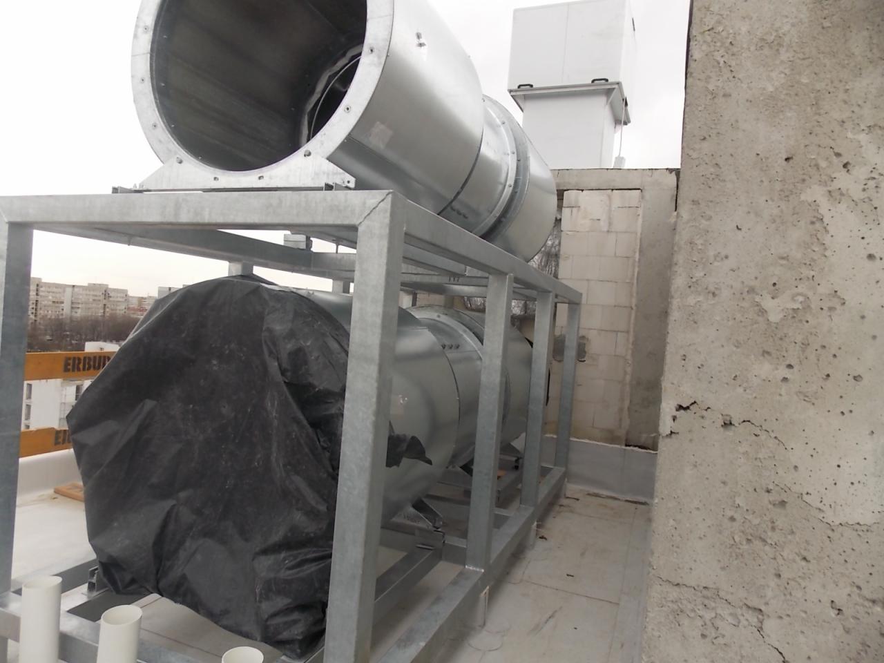 Instalacja przemysłowa na dachu: system rur wentylacyjnych z metalową konstrukcją wsporczą, jedna sekcja przykryta czarną folią, w tle budynki mieszkalne.