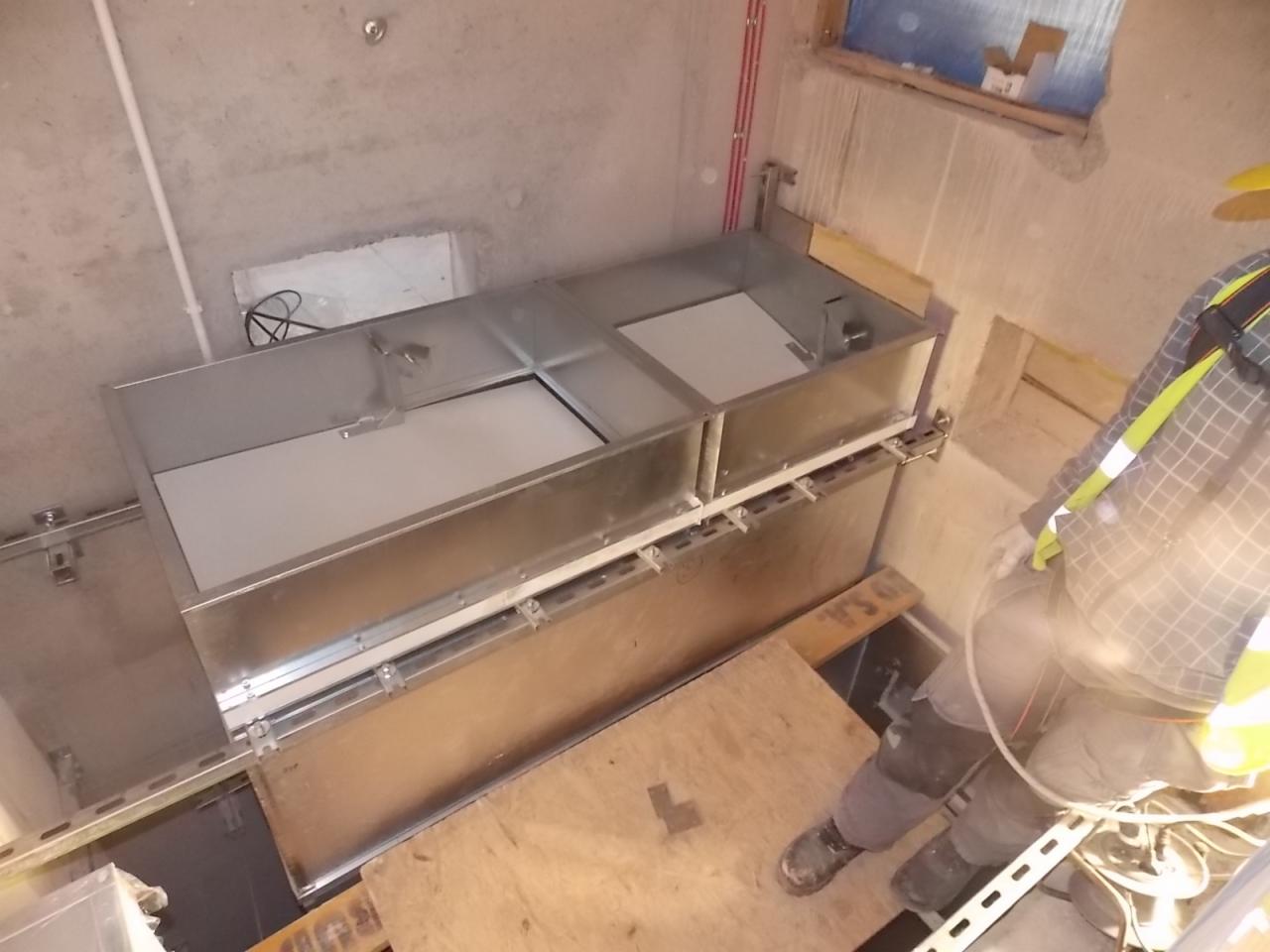 Metalowa konstrukcja z białymi panelami w surowym wnętrzu, częściowo widoczna osoba w odblaskowej kamizelce, prawdopodobnie w trakcie montażu.