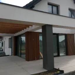 Nowoczesny dom z białymi ścianami i drewnianą pergolą nad tarasem wyłożonym jasnymi płytami, widoczne przesuwne okna i drewniane panele elewacyjne