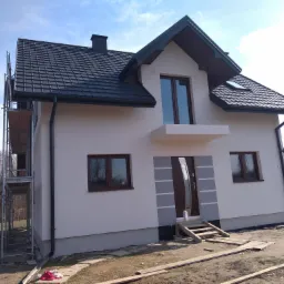 Nowy dom jednorodzinny z ocieploną, pomalowaną na biało elewacją, czarną dachówką i brązowymi oknami, z widocznymi rusztowaniami i materiałami budowlanymi na posesji.