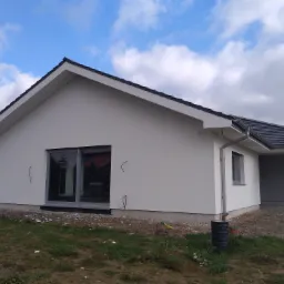 Nowoczesny, parterowy dom z białą elewacją, szarymi oknami i ciemną dachówką, widoczny fragment ogrodu z ziemią i trawą, przewody elektryczne wystające ze ściany, pochmurne niebo w tle.