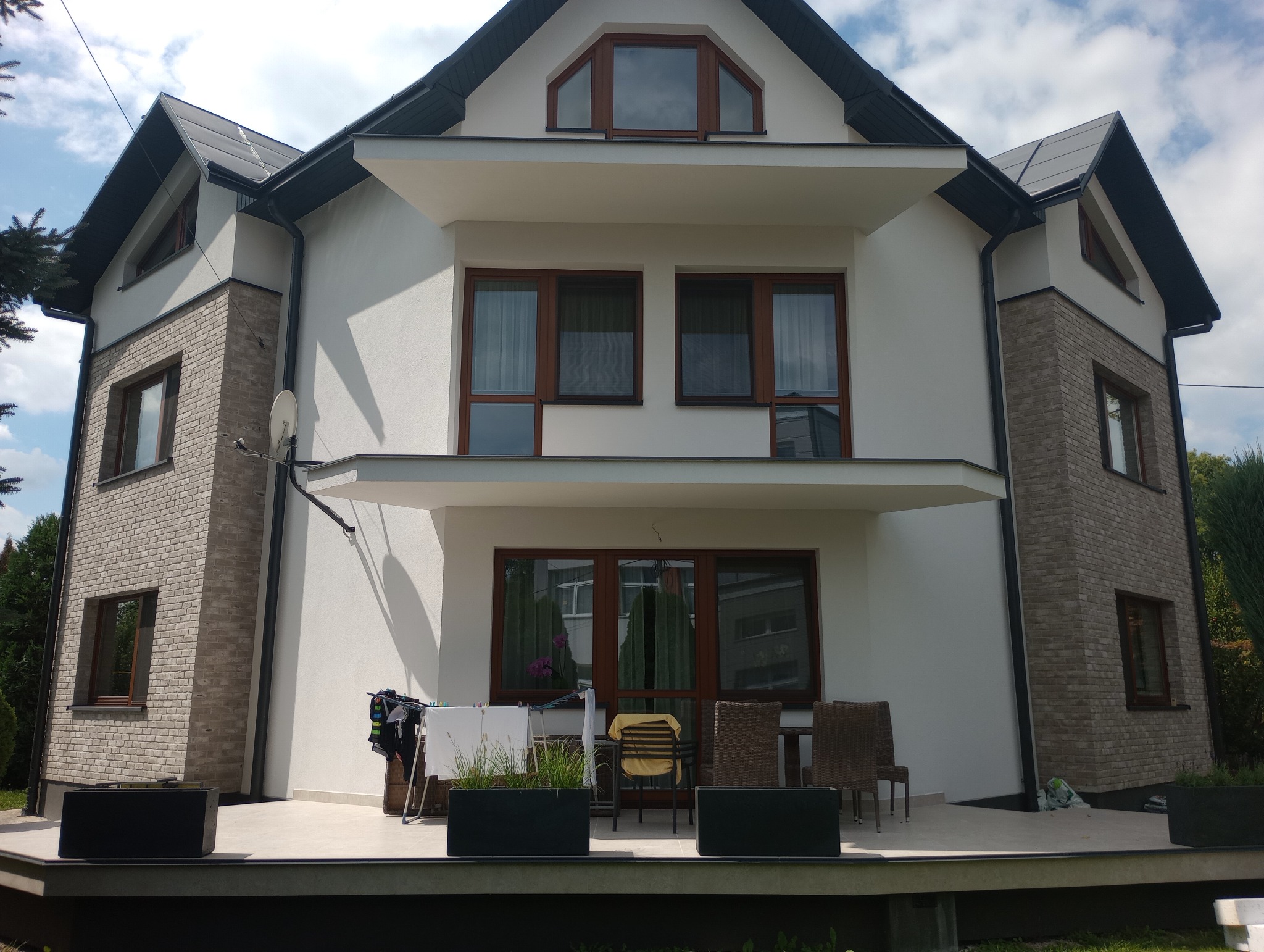 Nowoczesny dom dwukondygnacyjny z białymi ścianami i fragmentami elewacji z cegły, ciemne okna i dach, widoczne elementy zagospodarowania terenu, w tym taras z meblami i roślinami w donicach.