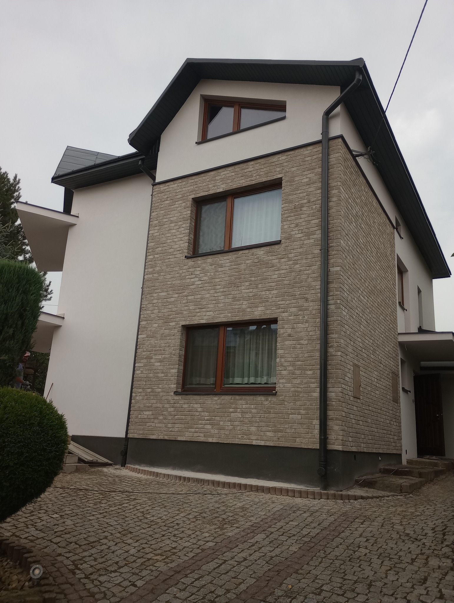 Dwukondygnacyjny dom z elewacją częściowo wykończoną jasną cegłą, z oknami w drewnianych ramach i ciemną dachówką, na tle pochmurnego nieba.